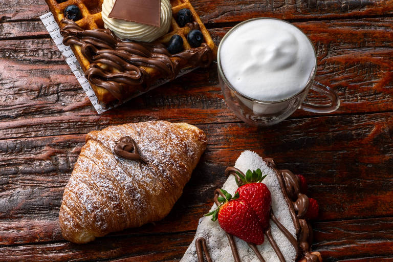 A imagem apresenta uma mesa de madeira com várias iguarias. No centro, há uma fatia de panqueca coberta com chocolate e morangos. Ao lado, um croissant polvilhado com açúcar de confeiteiro e um waffle decorado com creme e mirtilos, também coberto com chocolate. Há uma xícara de vidro com espuma de leite ao lado.