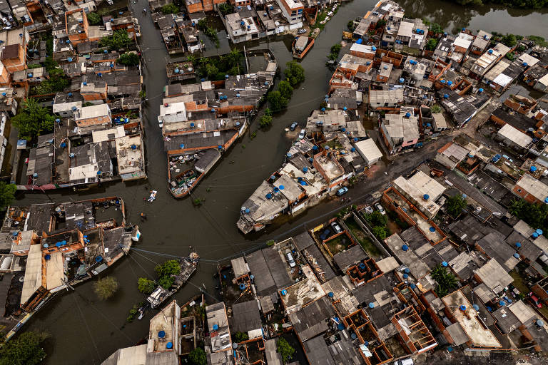 Imagem aérea de uma área urbana com casas dispostas em meio a ruas alagadas. As casas têm telhados de diferentes cores， principalmente em tons de azul e terracota， e estão cercadas por vegetação