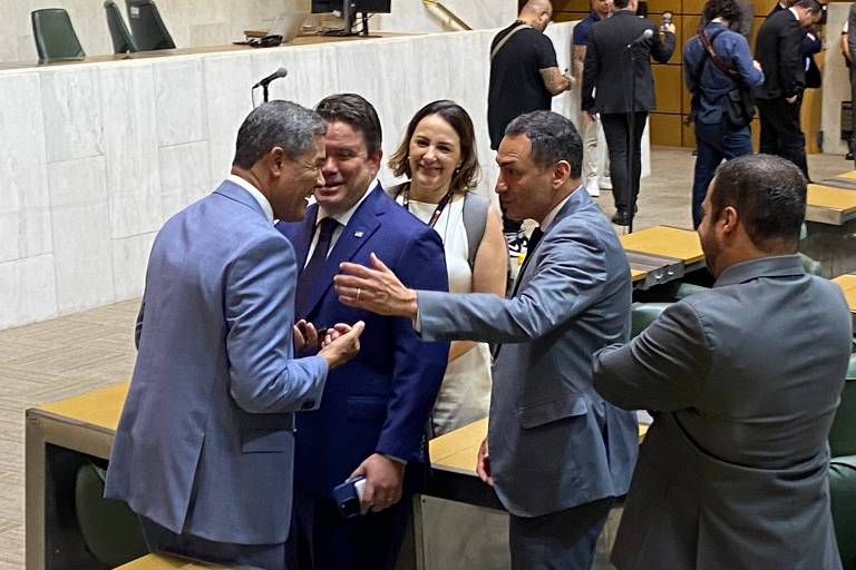 Um grupo de cinco pessoas interagindo em um ambiente político. Quatro homens estão em pé， conversando animadamente， enquanto uma mulher observa e sorri. O fundo mostra mesas e cadeiras típicas de um espaço legislativo. O ambiente é bem iluminado e formal.