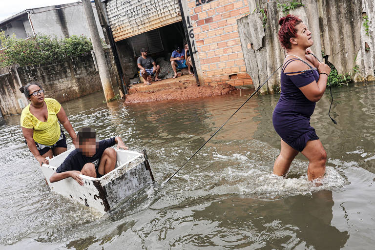 A imagem mostra uma cena de inundação, onde uma mulher está puxando uma caixa de metal com uma pessoa dentro, enquanto outra mulher a acompanha ao lado. O nível da água é alto, e ao fundo, algumas pessoas estão sentadas em uma área elevada. O ambiente parece ser uma rua inundada, com casas parcialmente submersas.