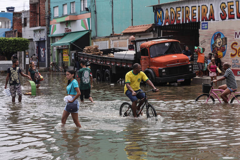 A imagem mostra uma rua inundada em uma área urbana， com várias pessoas caminhando e algumas montadas em bicicletas. Há um caminhão laranja estacionado ao fundo e lojas visíveis nas laterais. A água cobre grande parte da rua， refletindo o ambiente ao redor.