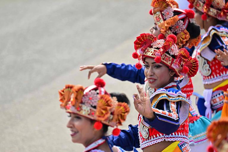 A imagem mostra um grupo de dançarinas em trajes tradicionais coloridos, com chapéus ornamentados e detalhes vibrantes. As dançarinas estão em movimento, com uma delas levantando a mão e sorrindo. O fundo é desfocado, sugerindo um ambiente de apresentação ou desfile.