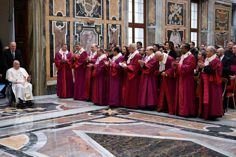 A imagem mostra um grupo de pessoas vestidas com túnicas vermelhas， formando um coro， em uma sala decorada com obras de arte. À esquerda， um homem sentado， que parece ser o Papa， observa o coro. O ambiente é elegante， com janelas grandes que permitem a entrada de luz natural