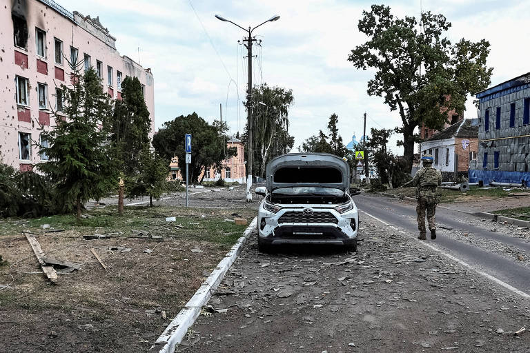 A imagem mostra uma rua em uma área urbana devastada， com um carro branco parado em primeiro plano. O chão está coberto de detritos e pedras， e há edifícios danificados ao fundo， com janelas quebradas e paredes em ruínas. Um soldado， vestido com uniforme militar， está caminhando ao lado do carro. Árvores e postes de luz estão visíveis， e o céu está nublado.