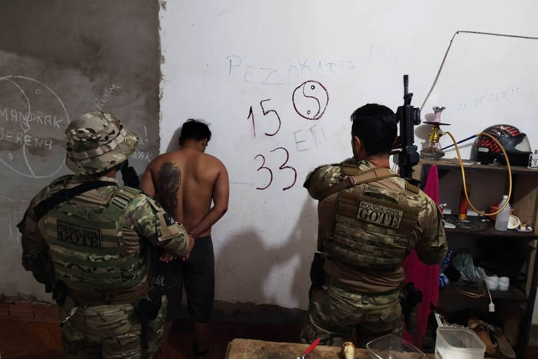 A imagem mostra dois homens em uniformes militares， um deles segurando uma arma， enquanto um terceiro homem， sem camisa， está de costas e preso. Na parede ao fundo， há números e símbolos escritos， incluindo 039;15039; e 039;33039;. O ambiente parece ser uma sala simples， com alguns objetos visíveis ao redor