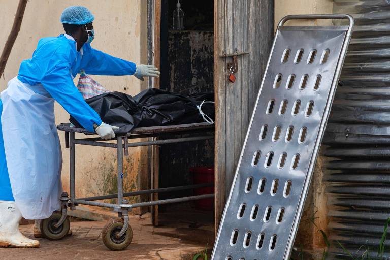 Um profissional de saúde， vestido com um traje de proteção azul e máscara， está empurrando uma maca com um corpo coberto por um lençol preto. Ao lado， há uma maca de metal com furos， encostada em uma parede. O ambiente parece ser uma área de atendimento médico ou hospitalar.