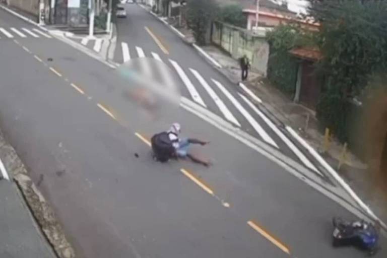 A imagem mostra uma cena de acidente de moto em uma rua. Um motociclista está caído no chão， enquanto a moto está deitada ao lado. A rua possui faixas de pedestres e árvores ao fundo. Não há veículos visíveis na imagem.