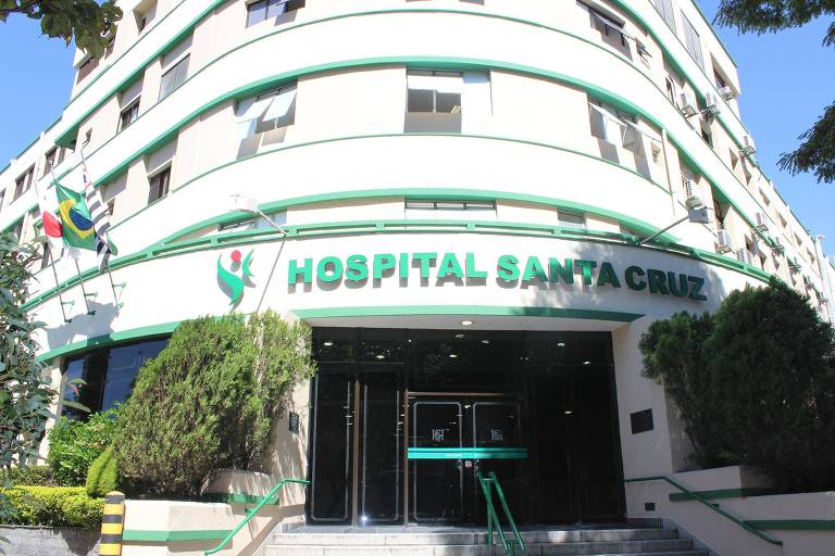 Imagem mostra a fachada do hospital. É um prédio de esquina com porta de vidro e tem o letreiro do hospital com sua logomarca em verde acima desta porta. Há canteiros com arbustos dos dois lados da porta