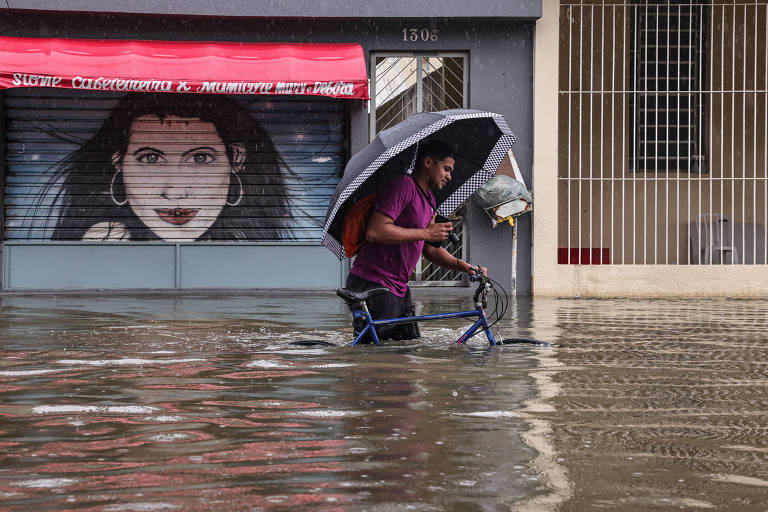 O homem está com as pernas mergulhadas na água enquanto caminha empurrando uma bicicleta com a mão esquer e segurando um guarda-chuva com a mão direita; ao fundo， a porta de um estabelecimento fechado tem o desenho do rosto de uma mulher branca de cabelos negros