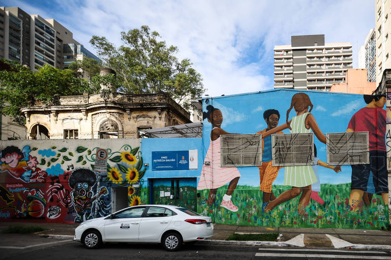 A imagem mostra uma cena urbana com murais coloridos em um lado e um edifício antigo em ruínas do outro. À esquerda， há um mural vibrante com figuras e elementos naturais， enquanto à direita， um mural retrata crianças brincando. Um carro branco está estacionado na rua em frente aos murais.