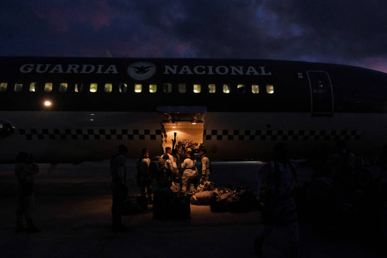 A imagem mostra um avião da Guarda Nacional com a porta de carga aberta， onde várias pessoas estão desembarcando. O ambiente é noturno， com nuvens escuras no céu. Algumas pessoas estão vestidas com uniformes e há bagagens no chão próximas ao avião.