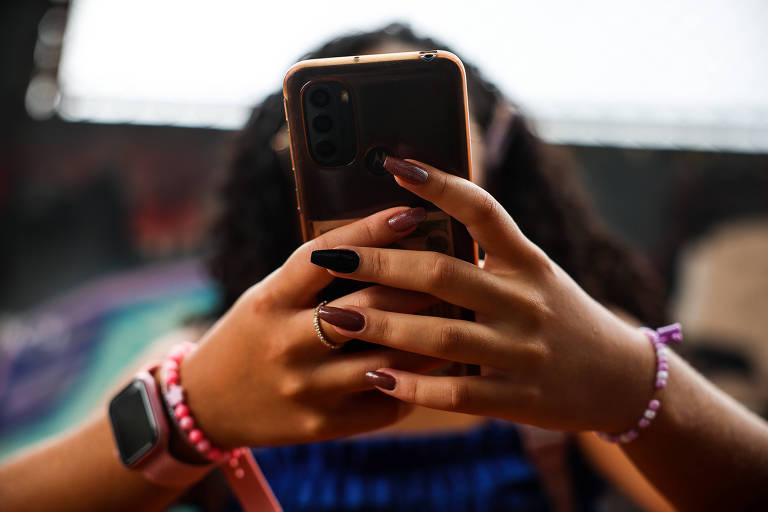 A imagem mostra uma pessoa segurando um celular com as duas mãos. As unhas estão pintadas em cores diferentes， uma mão tem unhas em tom nude e a outra em preto. A pessoa também usa pulseiras coloridas e um relógio inteligente no pulso. O fundo é desfocado， mas parece ter elementos coloridos.
