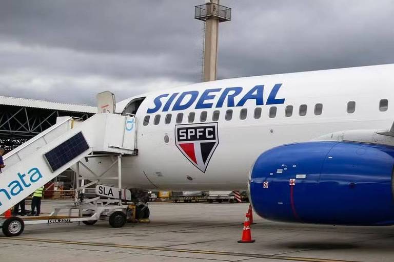 A imagem mostra um avião da companhia aérea Sideral estacionado em um aeroporto. O avião é branco com detalhes em azul e possui o logotipo do São Paulo Futebol Clube (SPFC) na fuselagem. Uma escada de embarque está posicionada ao lado do avião， e há um funcionário próximo a ela. O céu está nublado， com nuvens escuras， e ao fundo é possível ver uma torre de controle.