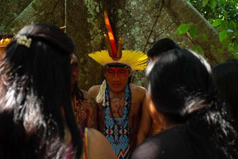 A imagem mostra um grupo de pessoas em uma cerimônia indígena. No centro, um homem com um cocar de penas amarelas e pintura facial vermelha está cercado por outros indivíduos. Ele usa um colar colorido e tem tatuagens visíveis no corpo. O fundo é composto por uma grande árvore e vegetação densa.