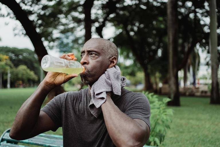 Um homem está bebendo de uma garrafa de bebida esportiva em um parque. Ele tem pele escura e cabelo grisalho， e está usando uma camiseta cinza. Um toalha está pendurada em seu pescoço. Ao fundo， há árvores e um gramado verde.