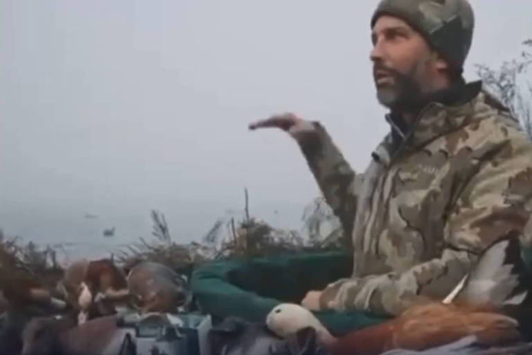 Um homem com um casaco camuflado está sentado em um ambiente natural， aparentemente em uma área de caça. Ele gesticula com a mão direita enquanto fala. Ao seu redor， há várias decorações de animais， possivelmente iscas， e uma paisagem nebulosa ao fundo.