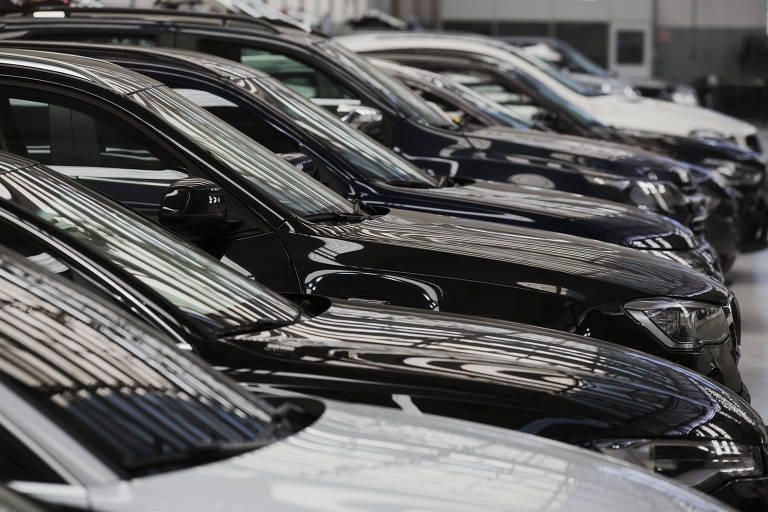 A imagem mostra uma fila de carros estacionados em um ambiente interno. Os veículos são predominantemente nas cores preta e branca， com reflexos visíveis na superfície dos carros. O ambiente parece ser uma concessionária ou um showroom， com iluminação artificial e um teto alto.