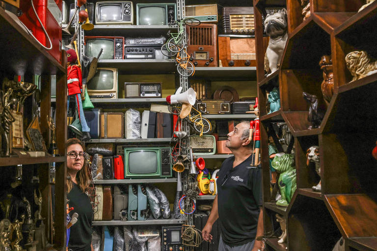 A imagem mostra um antiquário repleto de itens vintage, incluindo televisores antigos, rádios, e objetos decorativos. Duas pessoas estão no centro da imagem, uma mulher à esquerda e um homem à direita, ambos observando os itens ao redor. As prateleiras estão cheias de objetos variados, criando um ambiente de nostalgia.