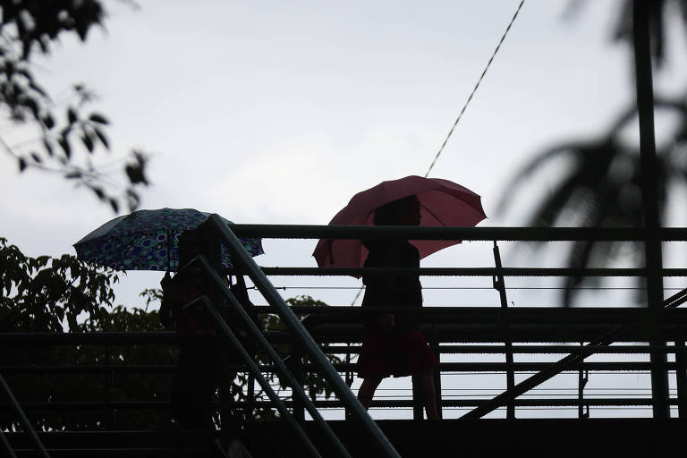 A imagem mostra duas pessoas caminhando em uma passarela， ambas segurando guarda-chuvas. Uma pessoa está com um guarda-chuva azul com estampas， enquanto a outra tem um guarda-chuva rosa. O fundo é de um céu nublado， criando um efeito de silhueta.