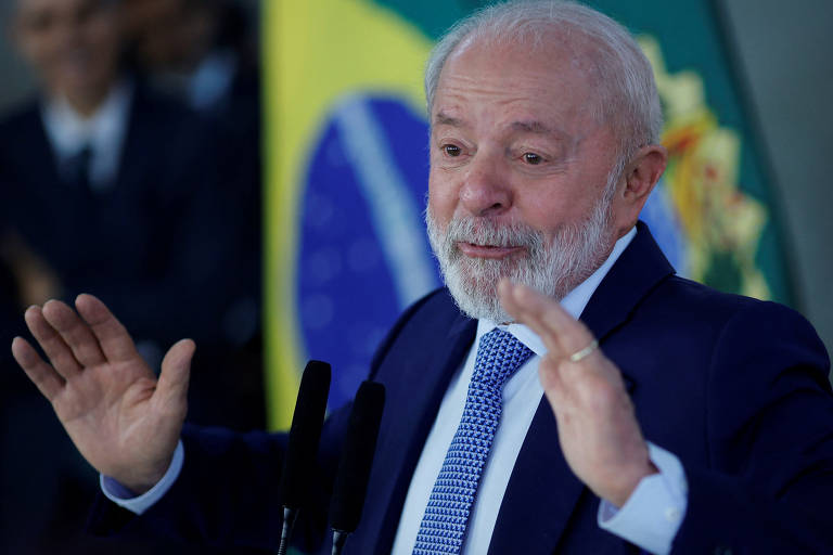 Lula, homem branco de cabelo grisalho, está de camisa social, terno escuro e gravata. Ao fundo, bandeira do Brasil. Ele está com as mãos à frente