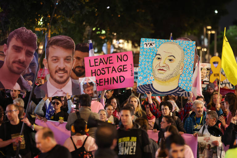 A imagem mostra um grande grupo de pessoas participando de um protesto à noite. Algumas pessoas seguram cartazes com rostos de indivíduos e mensagens， incluindo um que diz 039;LEAVE NO HOSTAGE BEHIND039;. O ambiente é iluminado por luzes e há uma atmosfera de mobilização social