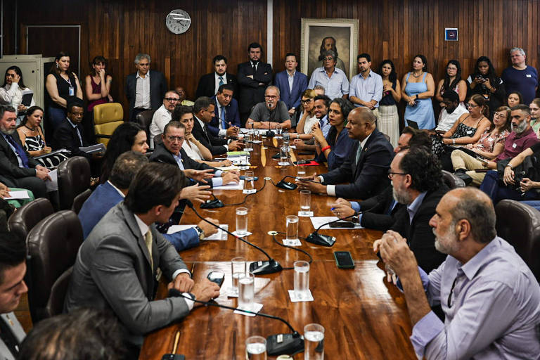 A imagem mostra uma reunião em uma sala com uma mesa longa e retangular. Há várias pessoas sentadas ao redor da mesa， algumas em trajes formais， outras em roupas casuais. No fundo， há uma parede de madeira e um quadro na parede. A sala está cheia de pessoas， incluindo observadores em pé. A mesa tem copos de água e alguns documentos visíveis.