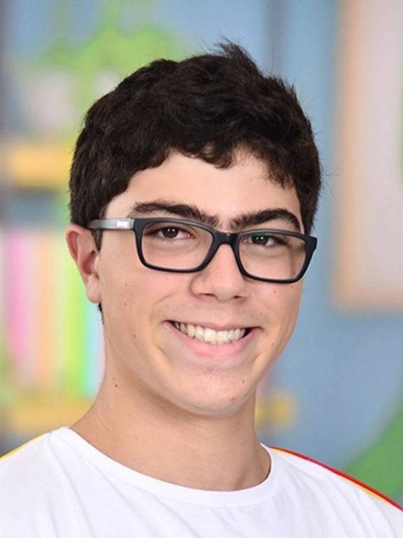 A imagem mostra um jovem sorridente com cabelo escuro e ondulado， usando óculos. Ele está vestindo uma camiseta branca e o fundo apresenta cores suaves e elementos que sugerem um ambiente escolar ou de aprendizado.