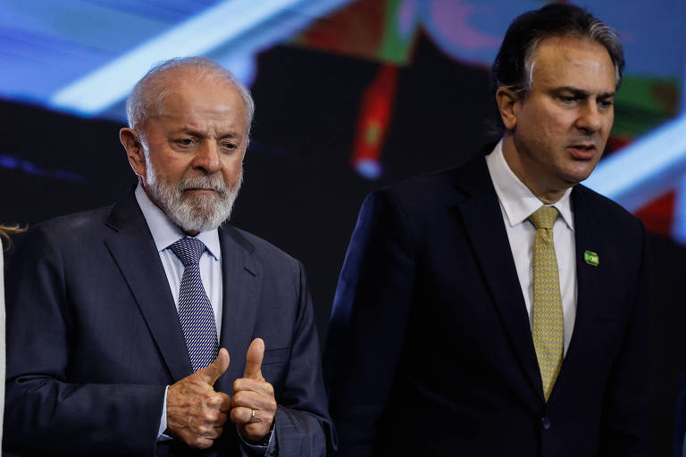 A imagem mostra dois homens em um evento. Lula, o homem à esquerda, tem cabelo grisalho e uma barba curta, vestindo um terno escuro e fazendo um gesto de positivo com o polegar. Camilo, à direita, está usando um terno escuro com uma gravata clara e parece estar em uma conversa. Ao fundo, há uma tela com cores variadas.