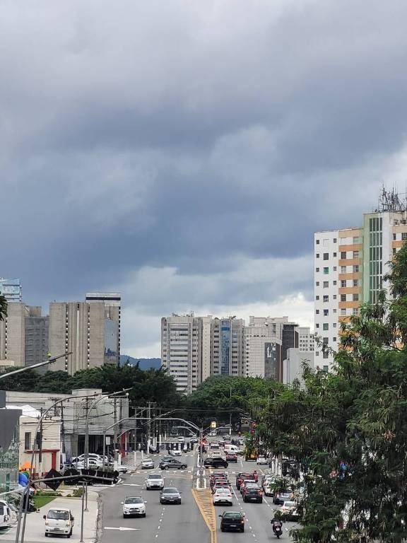 A imagem mostra uma vista de uma rua urbana com tráfego de veículos. Há prédios altos ao fundo e nuvens escuras no céu， sugerindo um clima nublado. A rua é larga e está cheia de carros em movimento， com algumas árvores visíveis nas laterais.