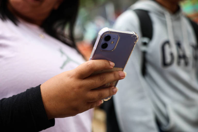 A imagem mostra uma estudante segurando um celular com uma capa transparente. A mão é visível， e a pessoa está usando uma blusa de manga longa preta. Ao fundo， há outra pessoa vestindo um moletom cinza com a palavra 039;GAP039;. O ambiente parece ser ao ar livre， com um fundo desfocado.