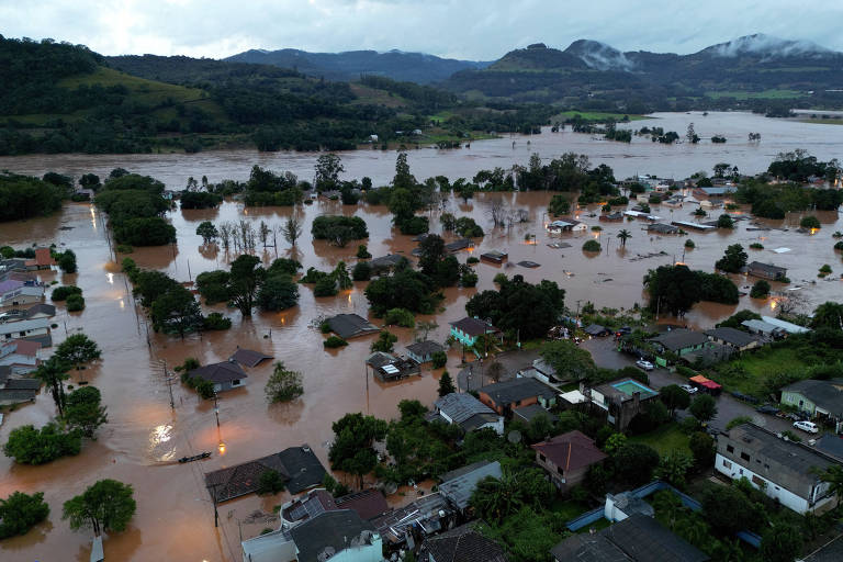 A imagem mostra uma vista aérea de uma área urbana afetada por inundações. Muitas casas estão parcialmente submersas em água marrom， com árvores e postes visíveis. O cenário é de um grande volume de água cobrindo ruas e propriedades， com montanhas ao fundo e um céu nublado.