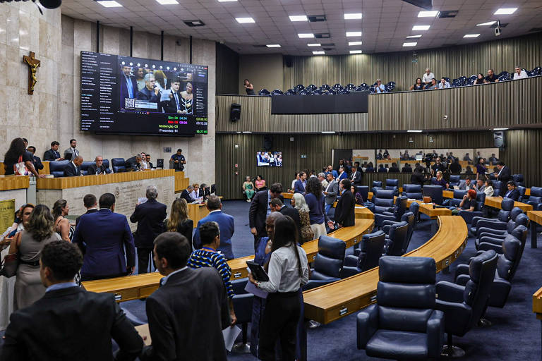 A imagem mostra o interior de um plenário legislativo, com várias pessoas em pé e sentadas. Há um grande painel eletrônico exibindo informações, e uma cruz está visível na parede. As cadeiras são dispostas em formato semicircular, e há mesas na parte frontal. O ambiente é bem iluminado e organizado, com um público observando as atividades.