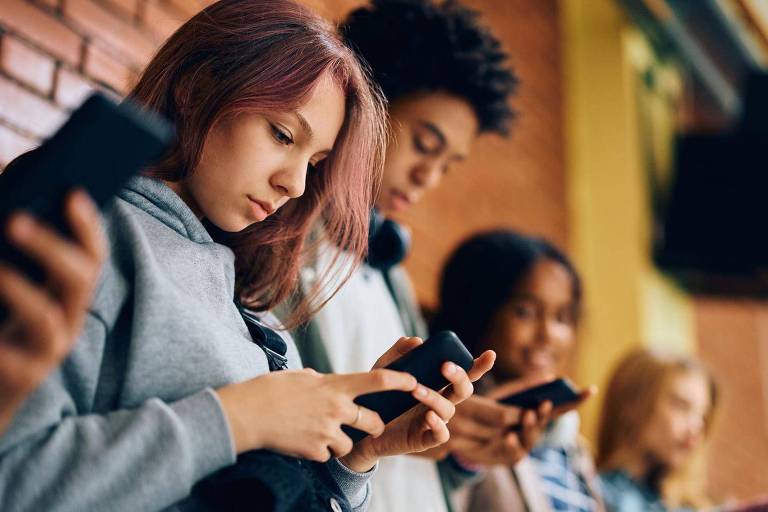 A imagem mostra um grupo de jovens em um ambiente interno， todos concentrados em seus celulares. Eles estão em pé， com as cabeças inclinadas para baixo， olhando para as telas. O fundo é de uma parede de tijolos e há um painel de madeira ao fundo. A iluminação é suave.