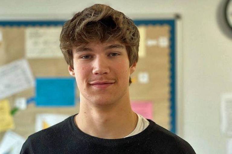 Um jovem com cabelo castanho claro e ondulado está posando para a foto. Ele usa uma camiseta preta e está em um ambiente escolar， com um fundo que mostra uma parede com papéis coloridos e um relógio. O jovem tem uma expressão neutra.