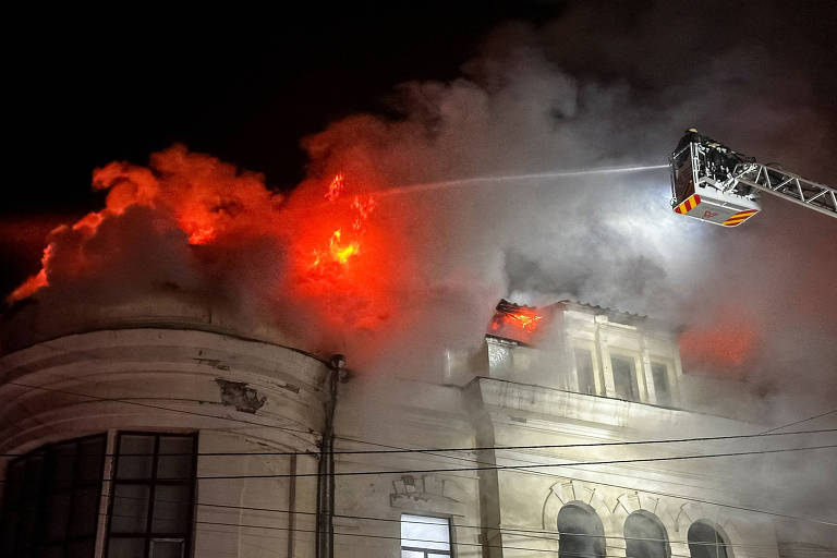 Bombeiros em uma escada jogam água sobre fogo em telhado de prédio branco antigo à noite