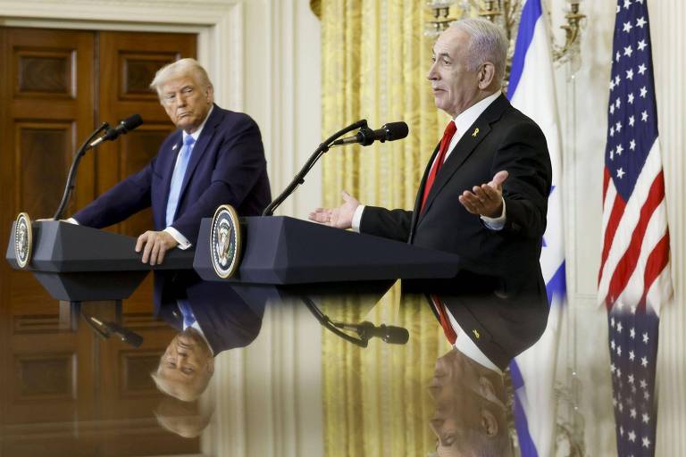 A imagem mostra dois homens em uma conferência de imprensa. À esquerda， um homem com cabelo loiro e terno escuro， e à direita， um homem com cabelo grisalho e terno vermelho. Ambos estão em frente a microfones， com bandeiras dos Estados Unidos e de Israel ao fundo. A mesa onde estão posicionados reflete suas imagens.