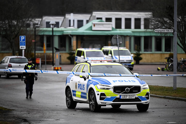 A imagem mostra um veículo policial estacionado em uma área com fita de isolamento. Um policial está em pé próximo ao carro， enquanto ao fundo há outros veículos e um edifício. O carro é um SUV da marca Volvo， com a palavra 039;POLIS039; visível em sua lateral. O ambiente parece ser urbano， com árvores e sinalização visíveis.

