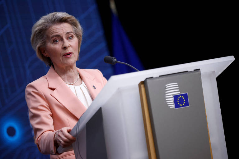 Uma mulher em um palanque de conferência， vestindo um blazer rosa claro， fala ao público. Ao fundo， há uma bandeira da União Europeia e um painel com um padrão geométrico. A mulher parece estar em um momento de discurso formal.
