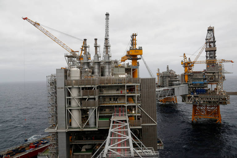 A imagem mostra várias plataformas de perfuração de petróleo no mar， com estruturas metálicas e guindastes visíveis. O céu está nublado， e as plataformas estão posicionadas próximas umas das outras， com uma delas em primeiro plano.
