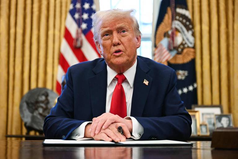 Um homem está sentado à mesa no Salão Oval， com as mãos cruzadas sobre um documento. Ele usa um terno escuro e uma gravata vermelha. Ao fundo， há cortinas douradas e a bandeira dos Estados Unidos. Também é visível um brasão presidencial e algumas fotos emolduradas na mesa.