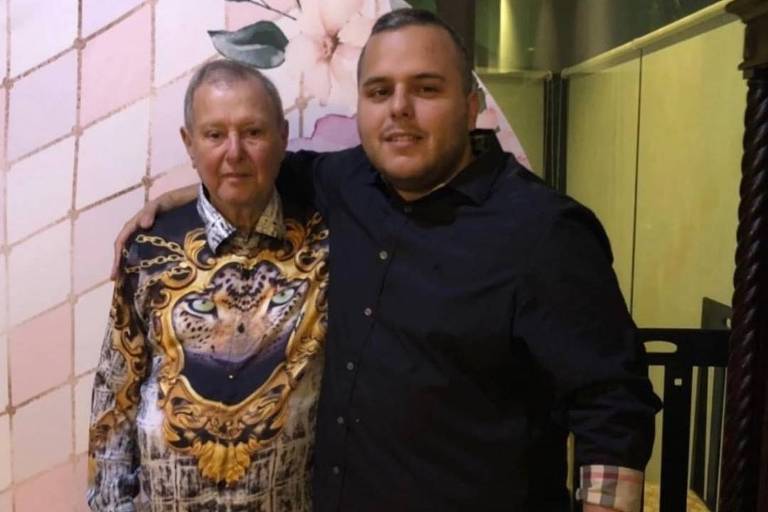 A imagem mostra dois homens posando juntos. O homem à esquerda está usando uma camisa colorida com estampas elaboradas e calças brancas. O homem à direita está vestido com uma camisa preta e calças escuras. Eles estão sorrindo e parecem estar em um ambiente festivo， com um fundo decorado com flores e padrões.