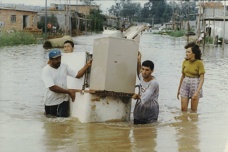 A imagem mostra um grupo de pessoas em uma área inundada, com água cobrindo a maior parte do chão. Quatro pessoas estão carregando um refrigerador, enquanto uma mulher observa. As casas ao fundo estão parcialmente submersas, e a cena retrata a dificuldade enfrentada pelos moradores devido à enchente.