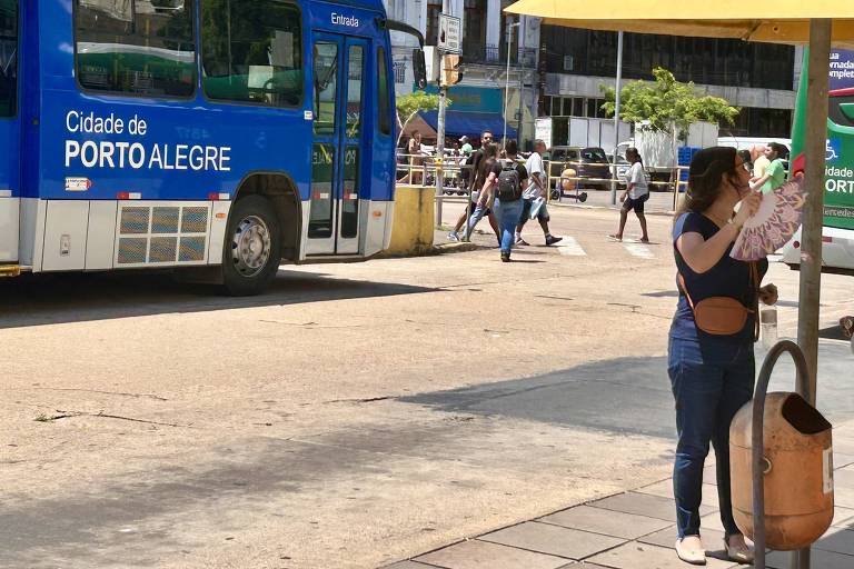 A imagem mostra uma parada de ônibus em Porto Alegre， com um ônibus azul ao fundo. Uma mulher está em pé ao lado da parada， interagindo com um dispositivo. O ambiente é urbano， com pessoas caminhando ao fundo e um quiosque amarelo na parte superior da imagem.