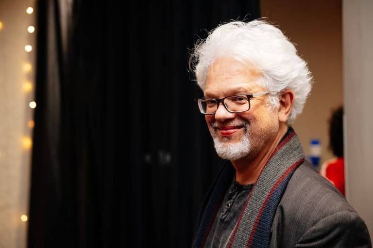 Um homem com cabelo grisalho e barba, usando óculos e uma jaqueta escura, está sorrindo enquanto olha para o lado. O fundo é desfocado, com uma parede clara e algumas luzes decorativas visíveis.