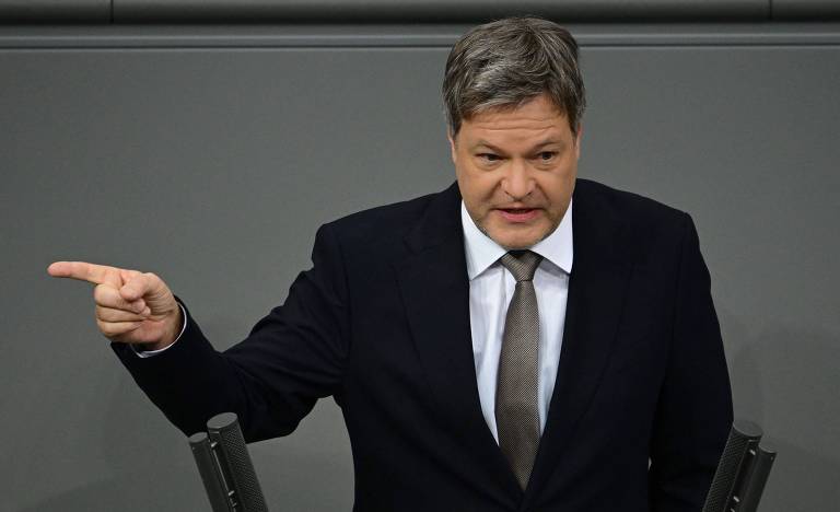 Um homem em um terno escuro está em pé， gesticulando com a mão direita enquanto fala em um ambiente que parece ser uma câmara legislativa. Ele tem cabelo curto e grisalho e parece estar enfatizando um ponto em sua fala.