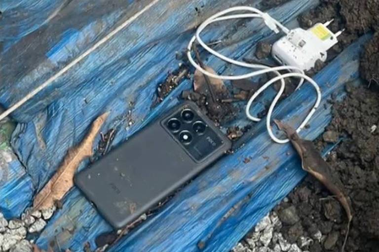 A imagem mostra um celular preto deitado sobre uma lona azul， ao lado de um carregador conectado a uma tomada. O fundo é composto por pedras pequenas e terra， com algumas folhas secas visíveis.