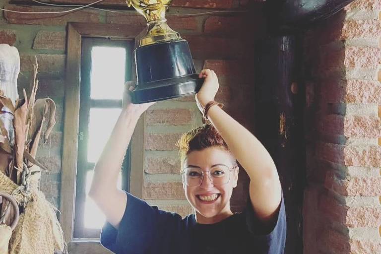 Uma pessoa está levantando um troféu dourado acima da cabeça em um ambiente interno. O troféu é grande e possui uma base preta. A pessoa está sorrindo e vestindo uma camiseta escura. Ao fundo， há uma parede de tijolos e uma janela com luz natural entrando.