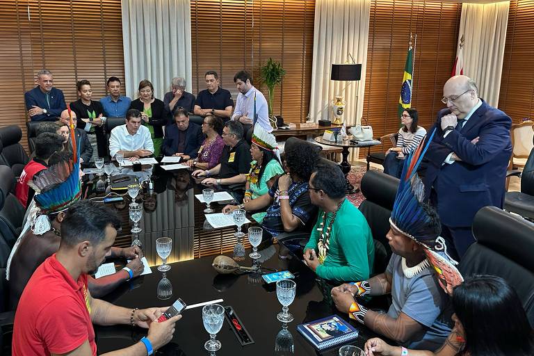 A imagem mostra uma reunião em um ambiente corporativo. Há várias pessoas sentadas ao redor de uma mesa retangular， com copos de vidro e materiais de escrita à frente. Algumas pessoas estão em pé， conversando. O ambiente é bem iluminado， com paredes de madeira e cortinas brancas. Uma bandeira do Brasil está visível ao fundo.