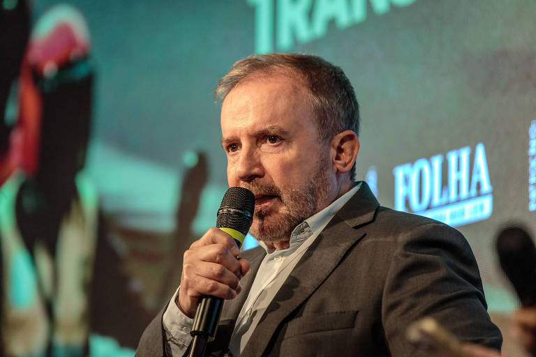 Um homem de cabelo curto e barba， vestido com um terno escuro， está falando ao microfone em um evento. Ao fundo， há uma tela com imagens e textos relacionados a um evento， incluindo o logotipo da Folha.
