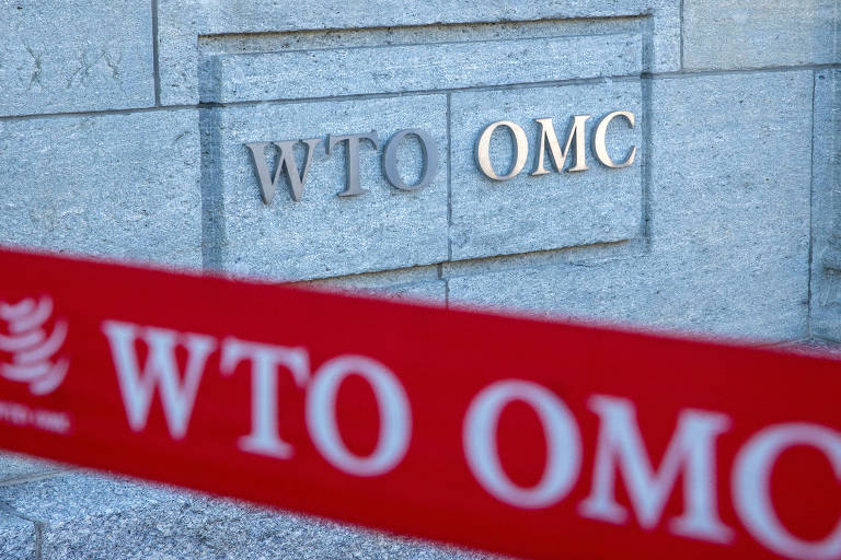 A imagem mostra o logotipo da Organização Mundial do Comércio (OMC)， com as letras 039;WTO039; e 039;OMC039; em destaque em uma placa de pedra. Na frente， há uma fita vermelha com as mesmas letras 039;WTO OMC039;.
